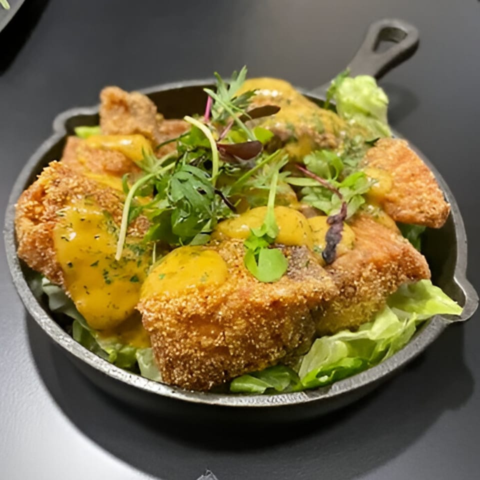 Crispy fried chicken pieces drizzled with yellow sauce and garnished with fresh greens in a skillet.