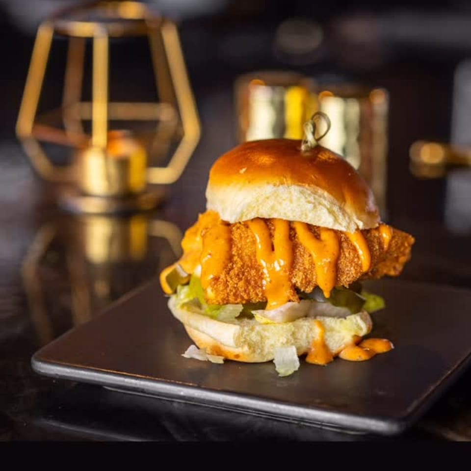 A crispy fried chicken slider with orange sauce, lettuce, and pickles on a dark plate.