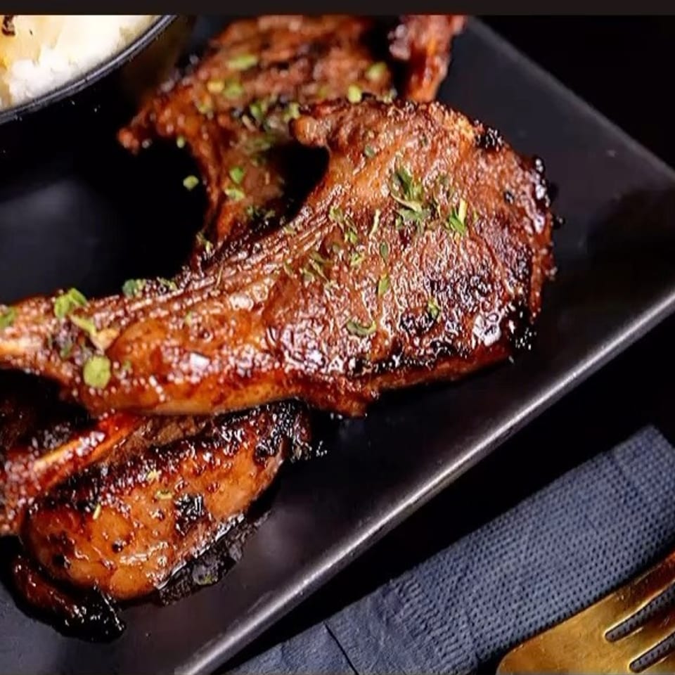 Soule To Soule's glazed lamb chops, garnished with herbs, on a black plate with a gold fork.